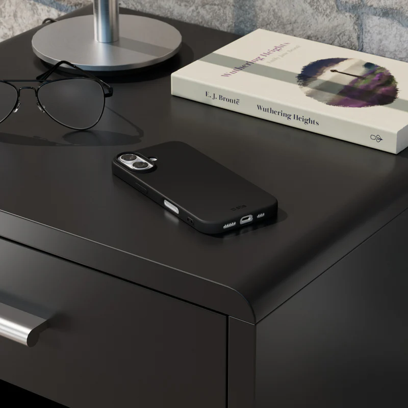 Ein schwarzes Telefon und eine Brille auf einem schwarzen Nachttisch. Ein Buch mit dem Titel 'Wuthering Heights' und eine Lampenbasis sind ebenfalls vorhanden.