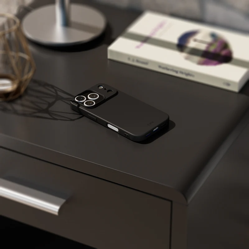 Ein schwarzes Smartphone mit Hülle liegt auf einem dunklen Nachttisch. Ein Buch und eine Lampe sind im Hintergrund.