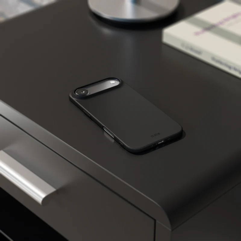 Ein schwarzes Smartphone mit Kameralinse liegt auf einem dunklen Tisch. Ein Buch und ein silberner Lampensockel sind im Hintergrund.