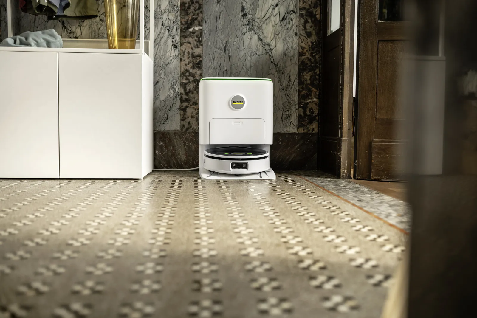 Biały odkurzacz robot stoi na wzorzystej podłodze. Biała szafka jest po lewej stronie. Drewniane drzwi są otwarte po prawej.