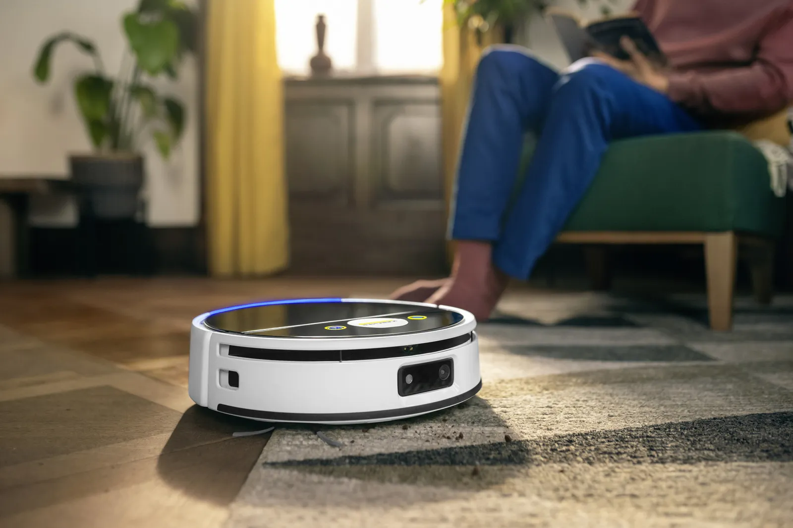 Robot odkurzający na dywanie. Osoba siedzi na zielonym krześle i czyta książkę. W tle meble i rośliny.