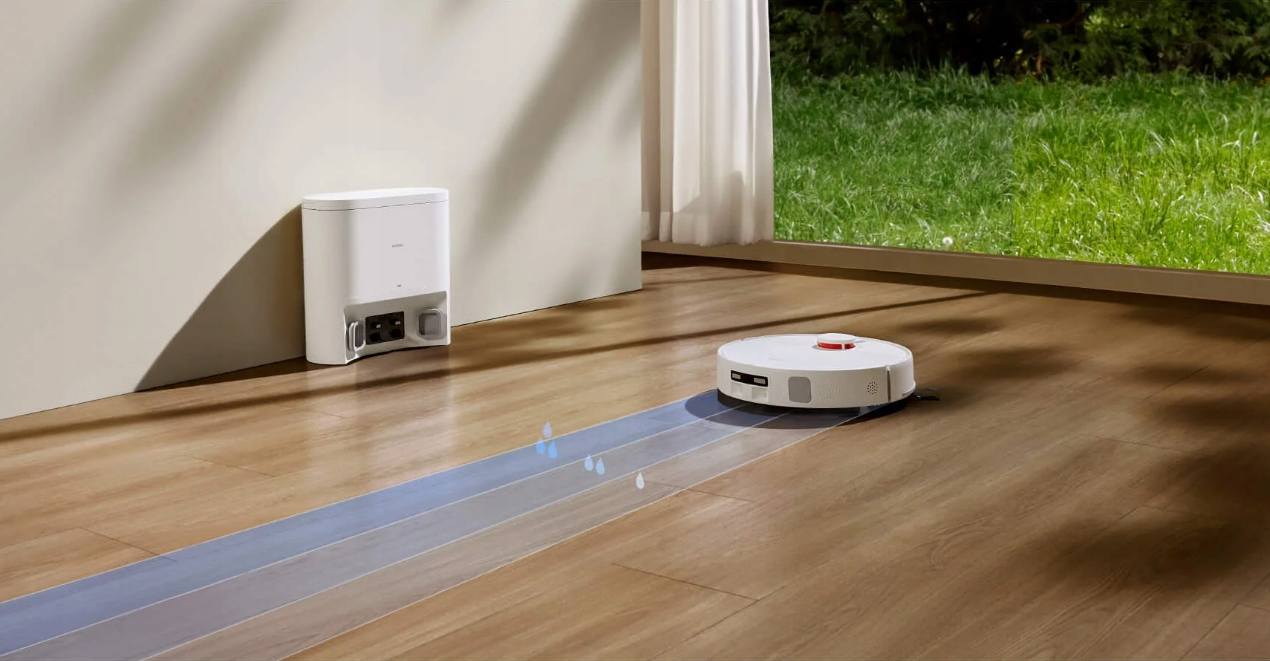 Biały robot odkurzający czyści drewnianą podłogę. Stacja ładująca jest po lewej stronie. Na zewnątrz widać zieloną trawę.