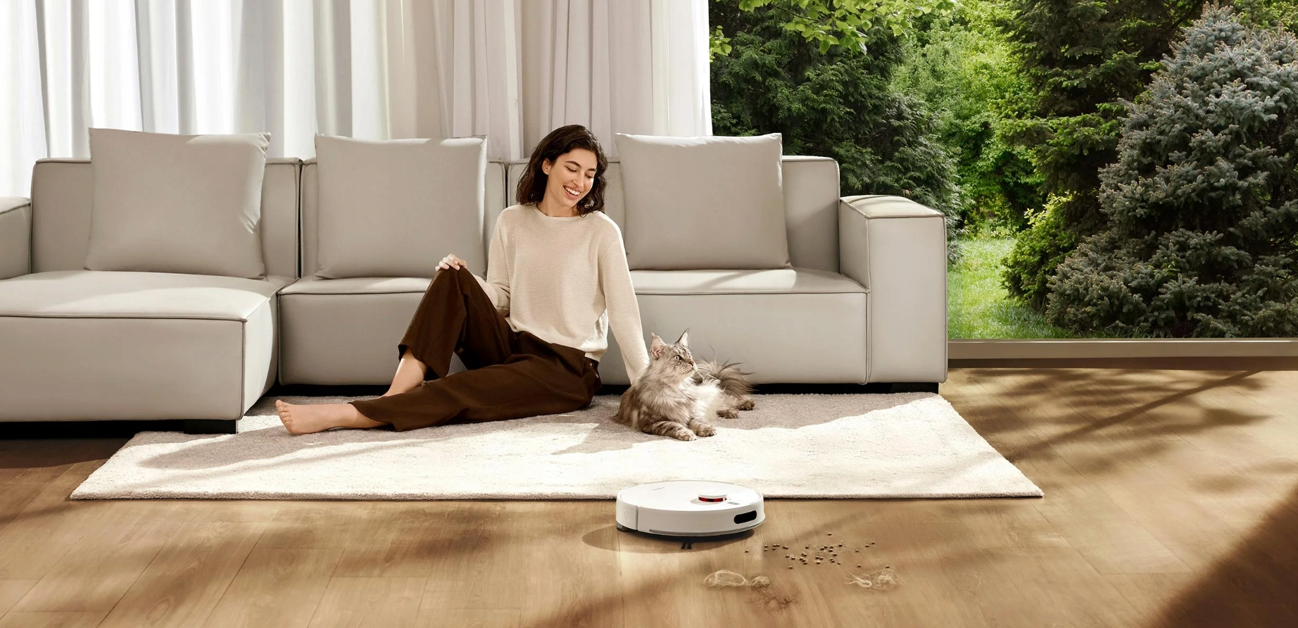 Kobieta z kotem siedzi obok białej sofy. Odkurzacz automatyczny na podłodze. Okno z drzewami w tle.
