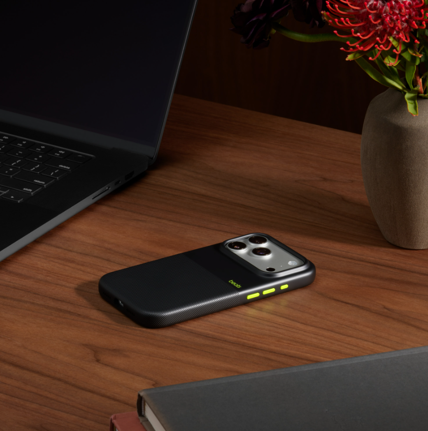 Siyah bir telefon kılıfı, bir dizüstü bilgisayar ve bir vazo çiçek ile ahşap bir masanın üzerinde duruyor. Kılıfın yeşil detayları ve dokulu bir sırtı var.