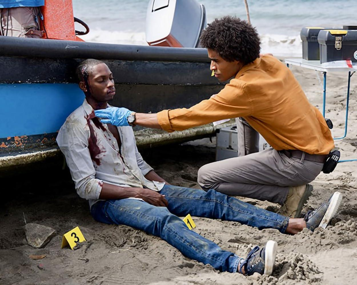 Ein Mann mit einer Kopfwunde liegt am Strand. Ein anderer, in einem orangefarbenen Hemd, untersucht ihn. Ein Boot und Markierungen sind in der Nähe.