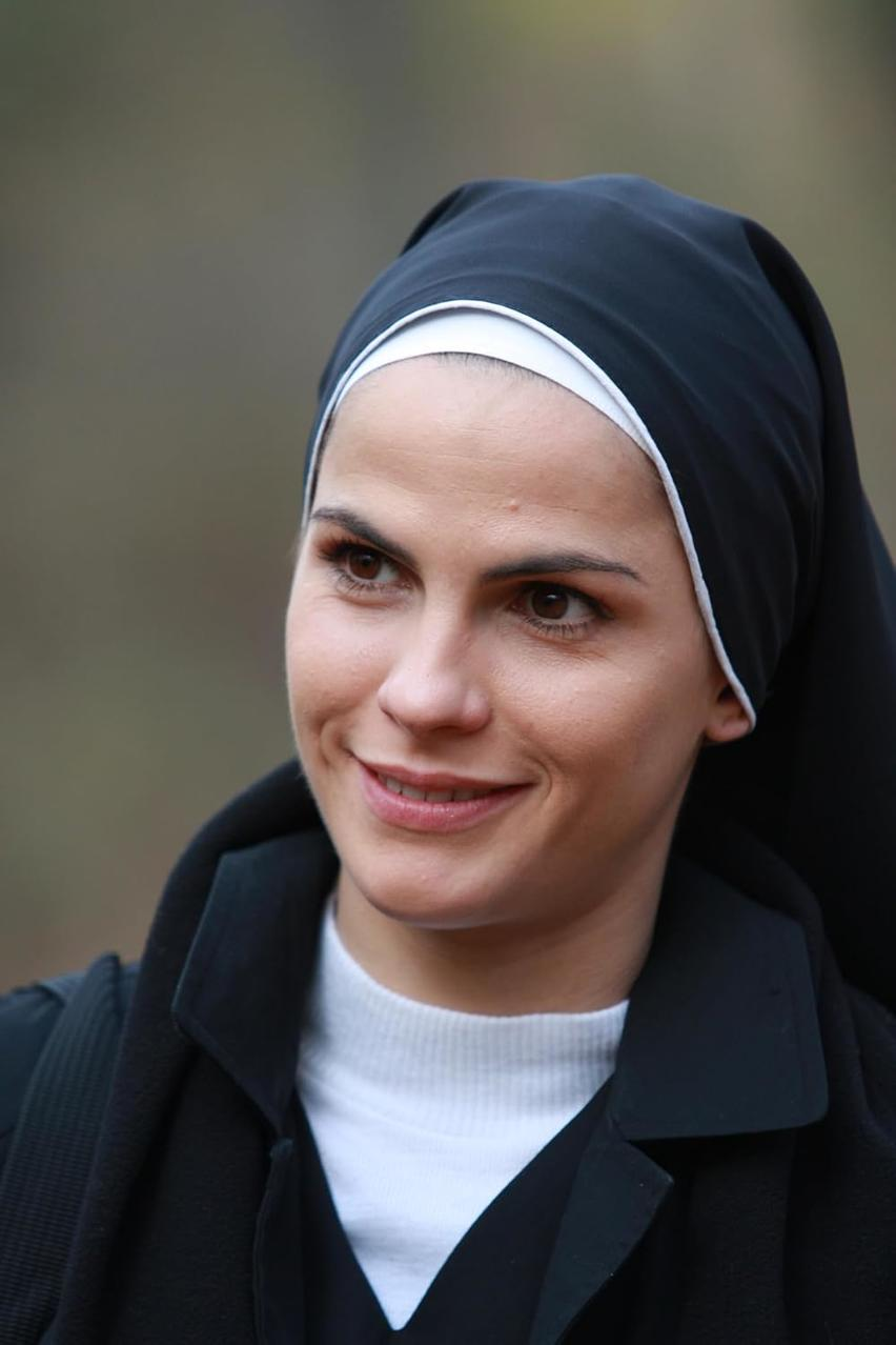 Eine Frau in Nonnentracht lächelt. Sie hat braune Augen und helle Haut. Der Hintergrund ist verschwommen mit Grün- und Brauntönen.