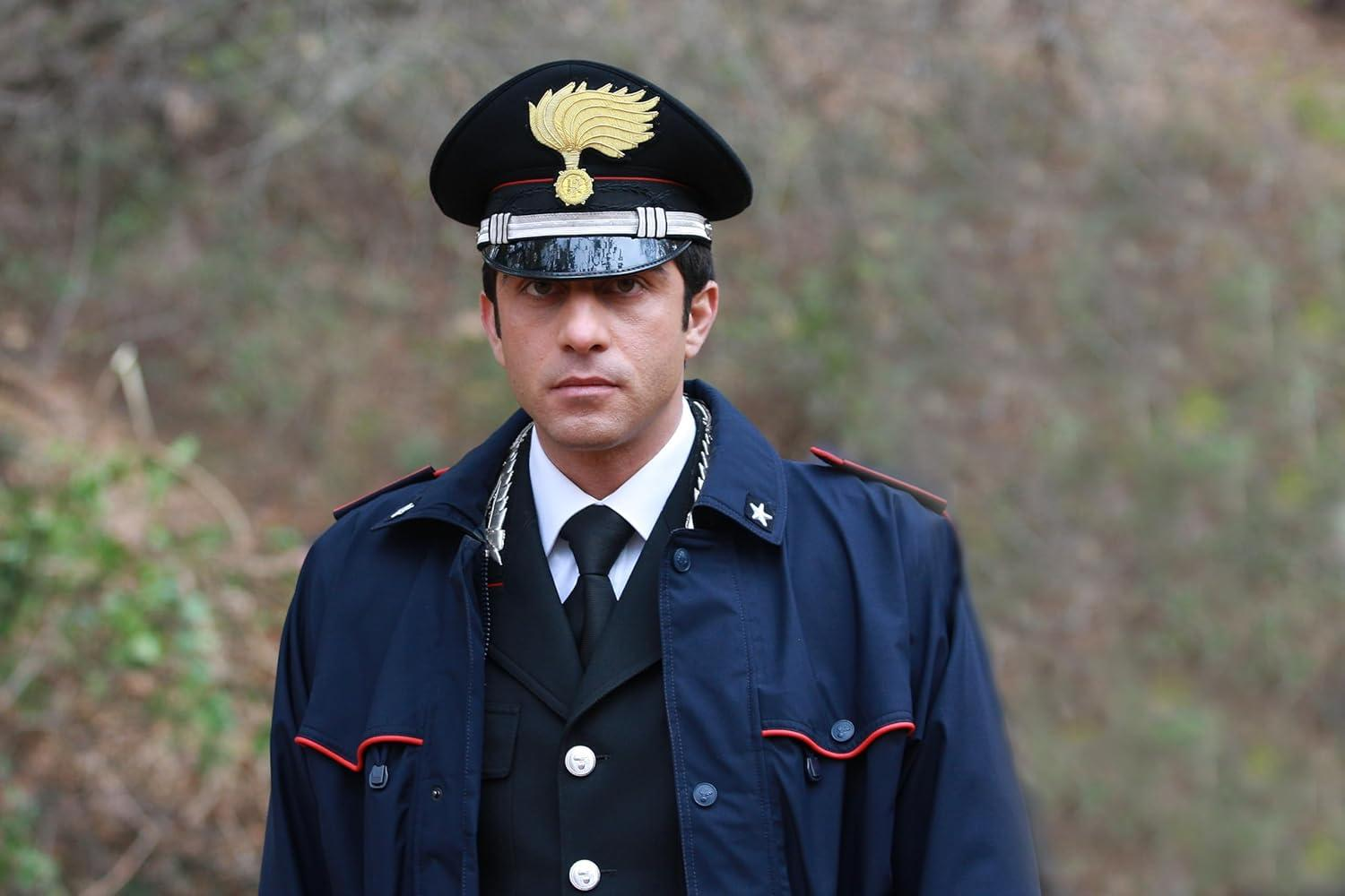 Ein Mann in dunkelblauer Polizeiuniform und Hut mit ernstem Gesichtsausdruck steht vor einem verschwommenen grünen Hintergrund.