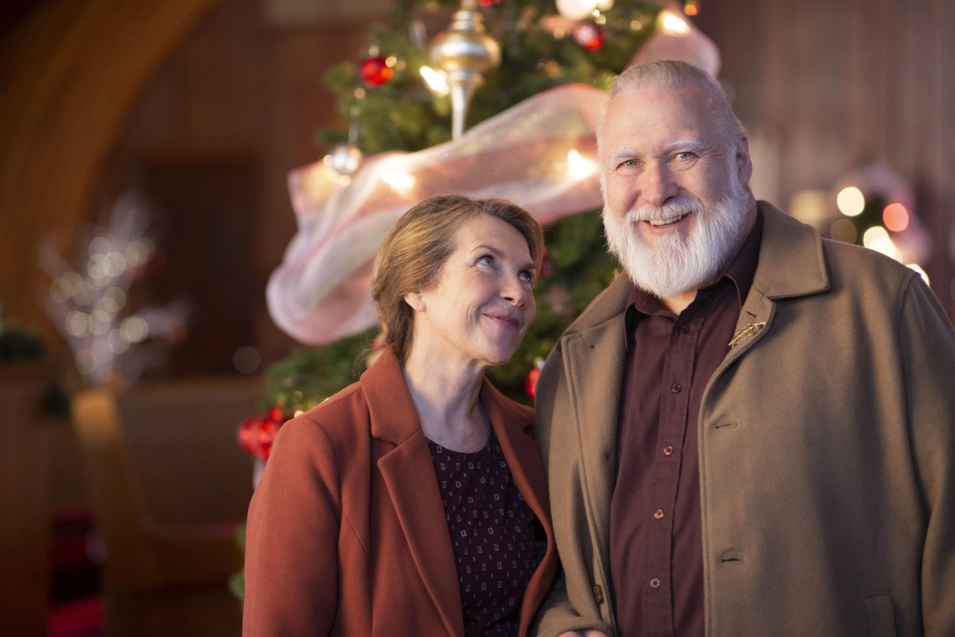 Ein lächelnder Mann und eine Frau stehen in der Nähe eines Weihnachtsbaums. Der Mann hat einen weißen Bart und die Frau schaut nach oben.