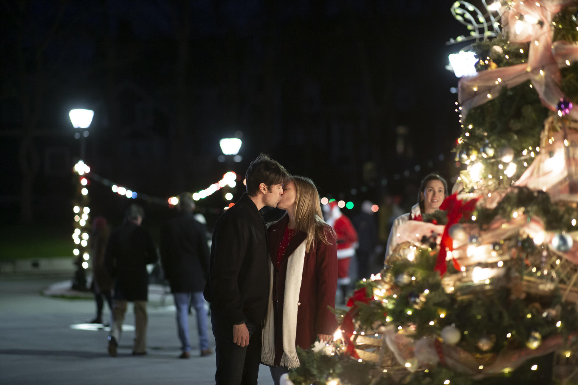 Ein Paar küsst sich in der Nähe eines geschmückten Weihnachtsbaums. Menschen und Lichter sind im Hintergrund.