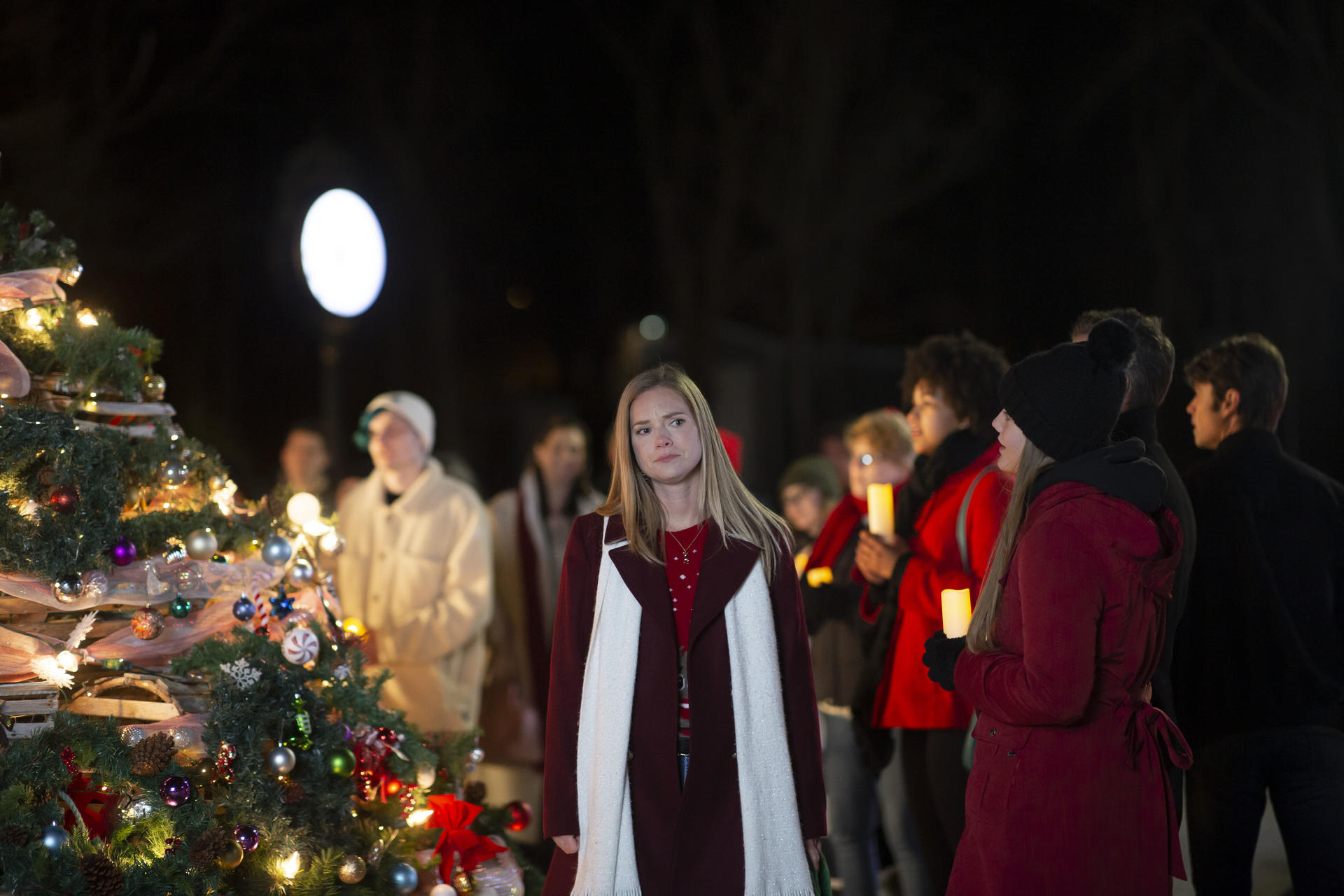 Eine Frau in einem bordeauxfarbenen Mantel und einem weißen Schal steht vor einem geschmückten Weihnachtsbaum. Menschen halten Kerzen. Die Szene spielt nachts.