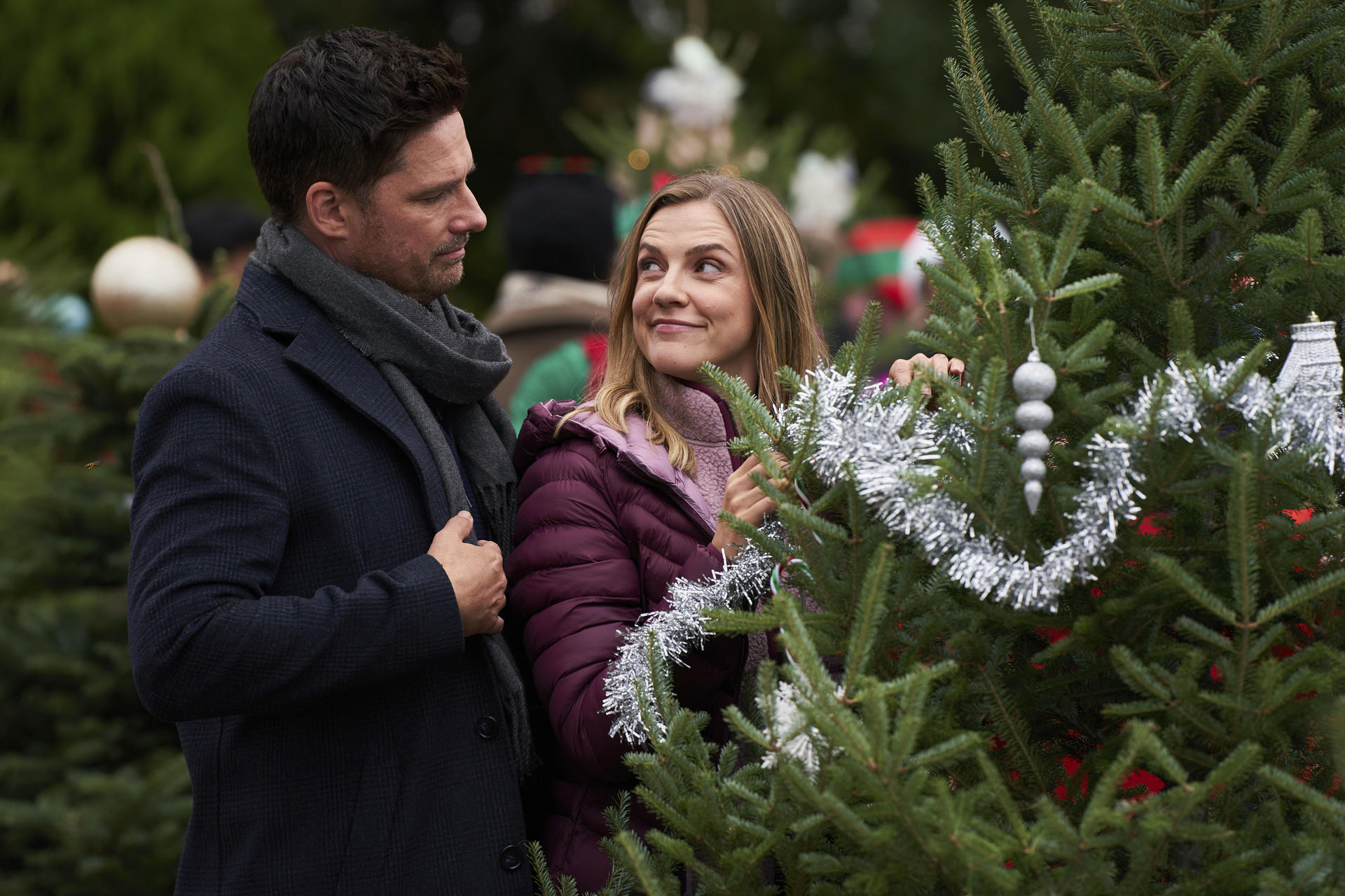Ein Mann und eine Frau stehen in der Nähe eines Weihnachtsbaums. Die Frau trägt eine lila Jacke. Der Mann trägt einen dunklen Mantel und Schal.