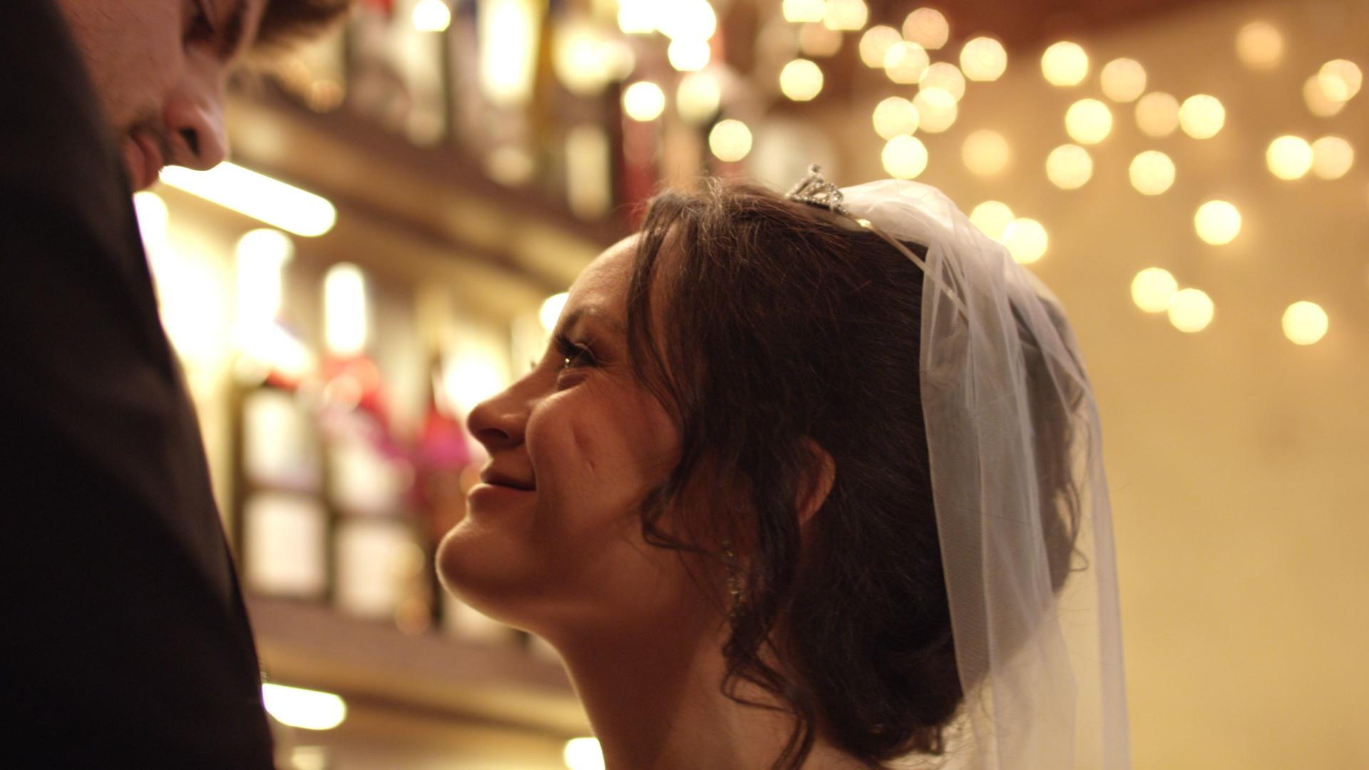 Eine Braut blickt lächelnd zu einem Mann auf. Sie trägt Schleier und Tiara. Weiche Lichter im Hintergrund deuten auf eine Hochzeit hin.