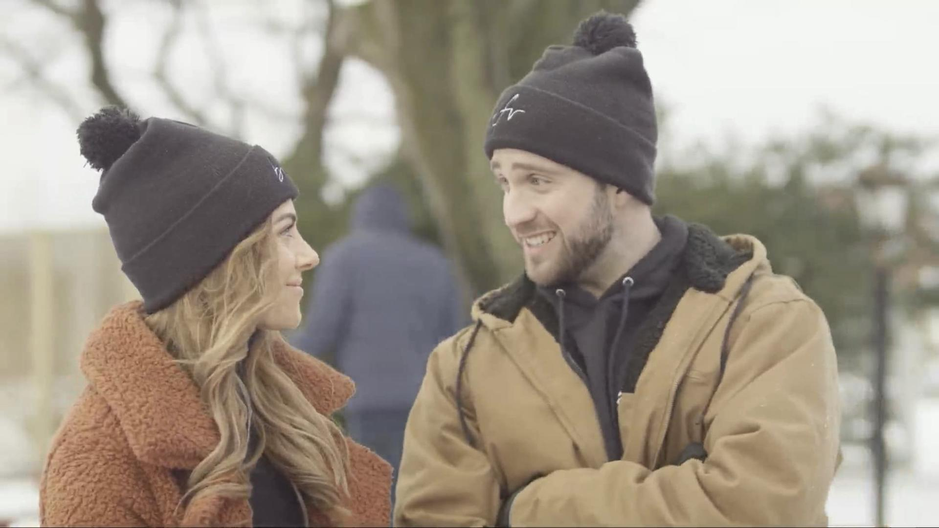 Ein Mann und eine Frau mit Mützen sehen sich an und lächeln. Beide tragen Winterjacken. Eine dritte Person ist im Hintergrund.