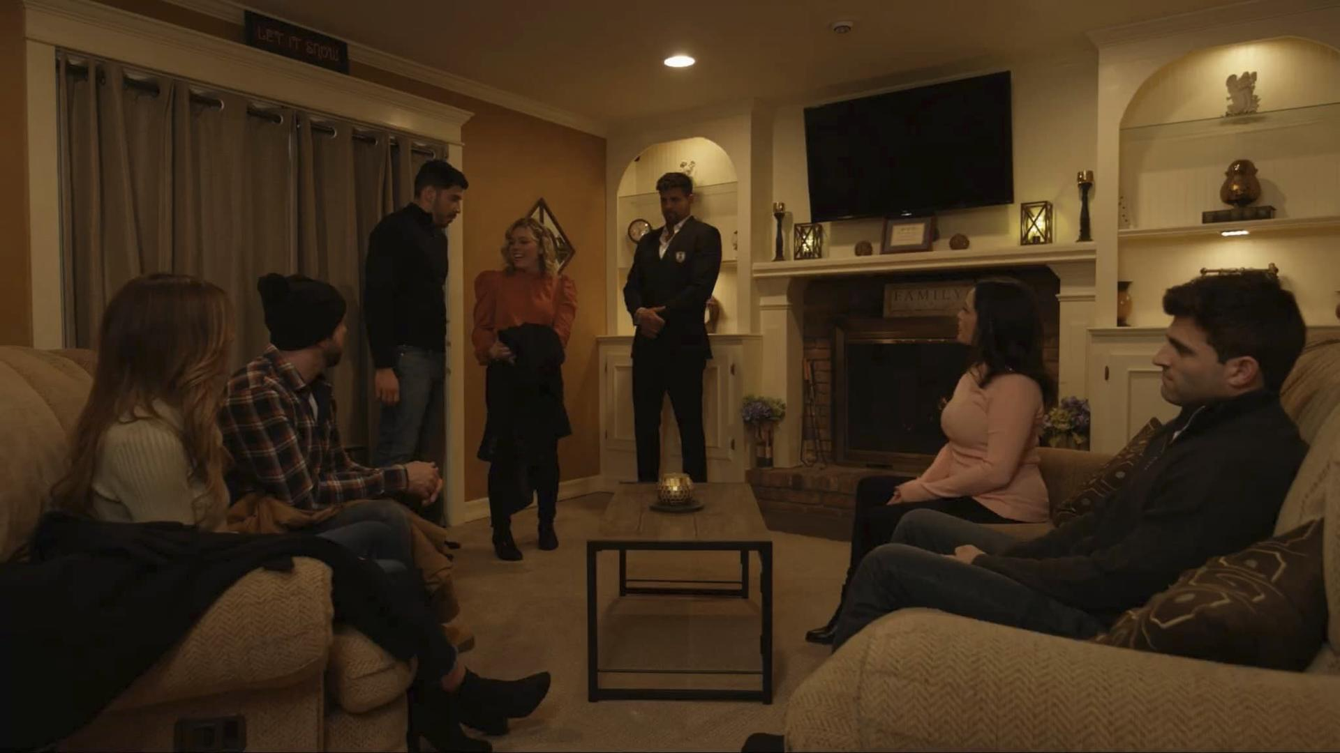 Eine Gruppe von Leuten in einem Wohnzimmer. Einige sitzen auf Sofas. Ein Mann im Anzug steht. Im Hintergrund befindet sich ein Kamin.