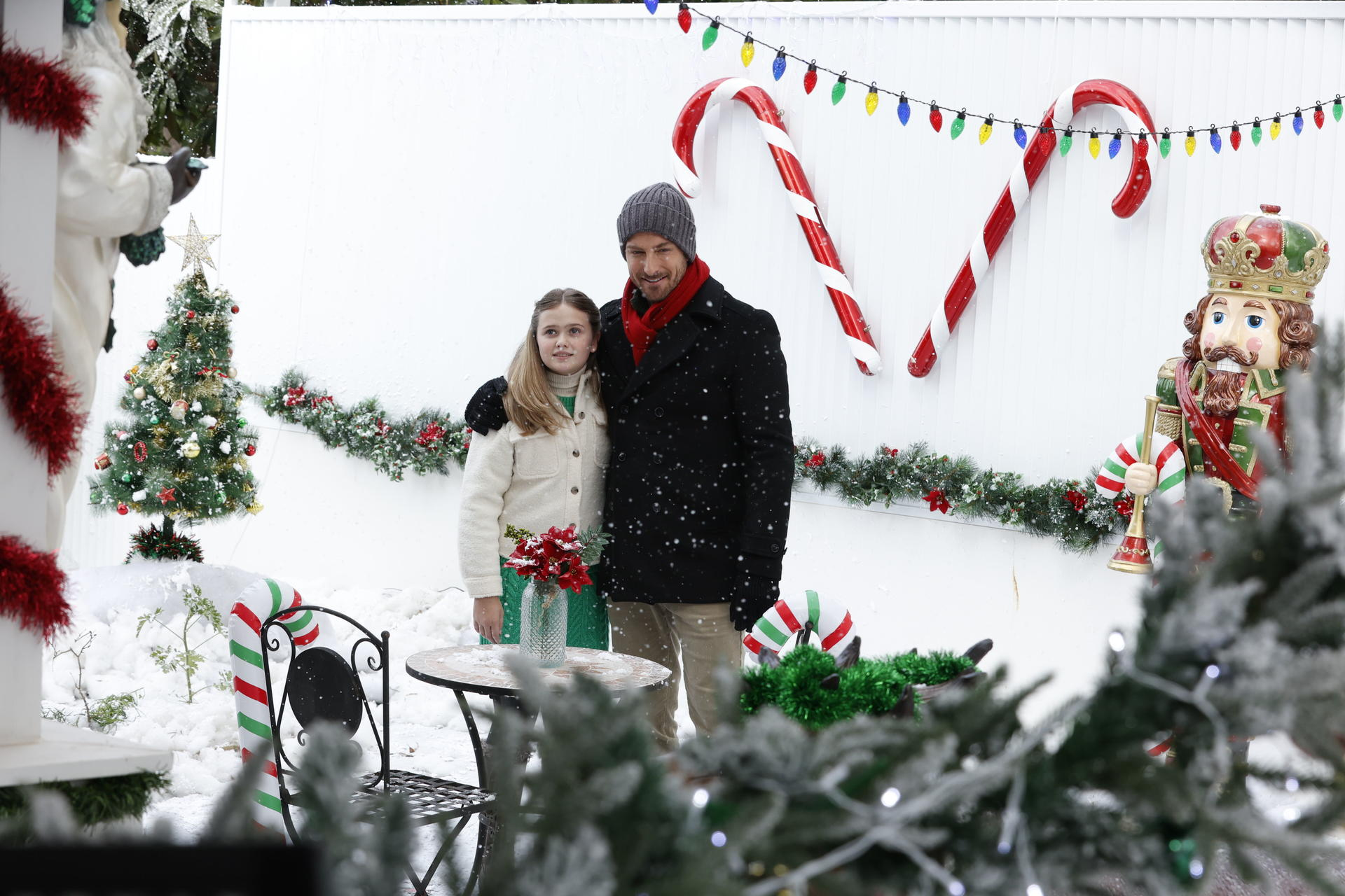 Ein Mann und ein Mädchen stehen vor einer weißen Wand mit Zuckerstangen und Weihnachtsbeleuchtung. Schnee fällt, festliche Dekorationen gibt es reichlich.