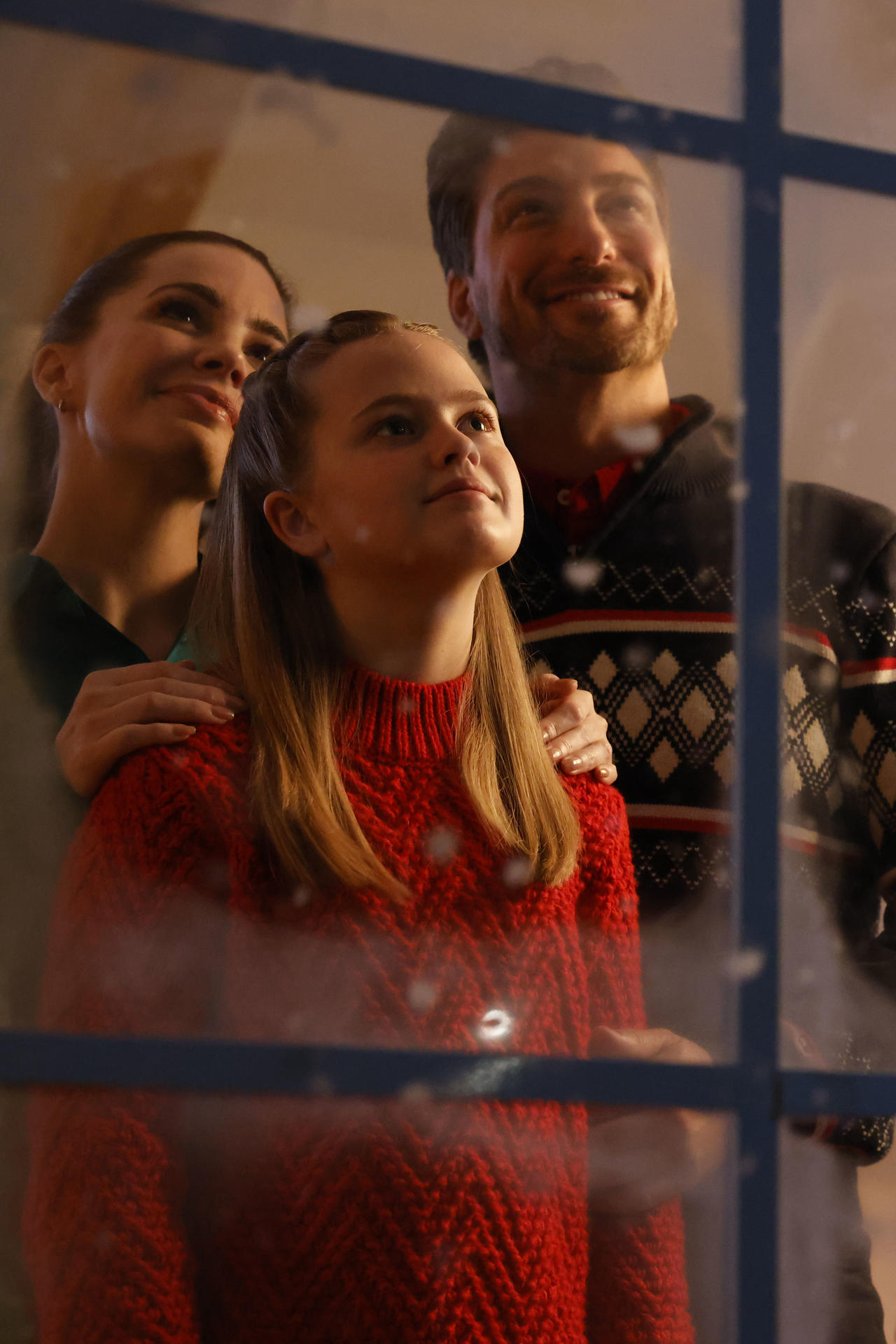 Eine Familie blickt durch ein Fenster nach oben. Ein Mädchen in einem roten Pullover steht vorne, die Eltern dahinter. Sie alle sehen glücklich aus.