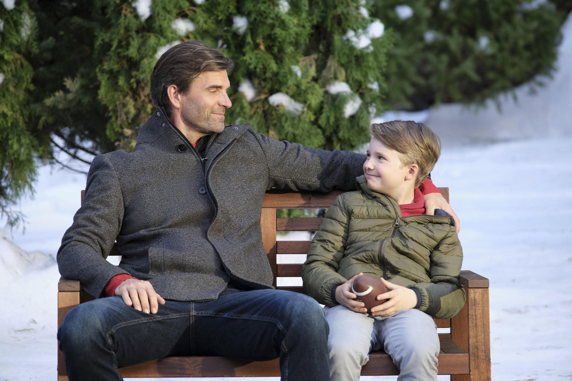Ein Mann und ein Junge sitzen auf einer Bank. Der Junge hält einen Football. Der Hintergrund umfasst Schnee und grüne Bäume.