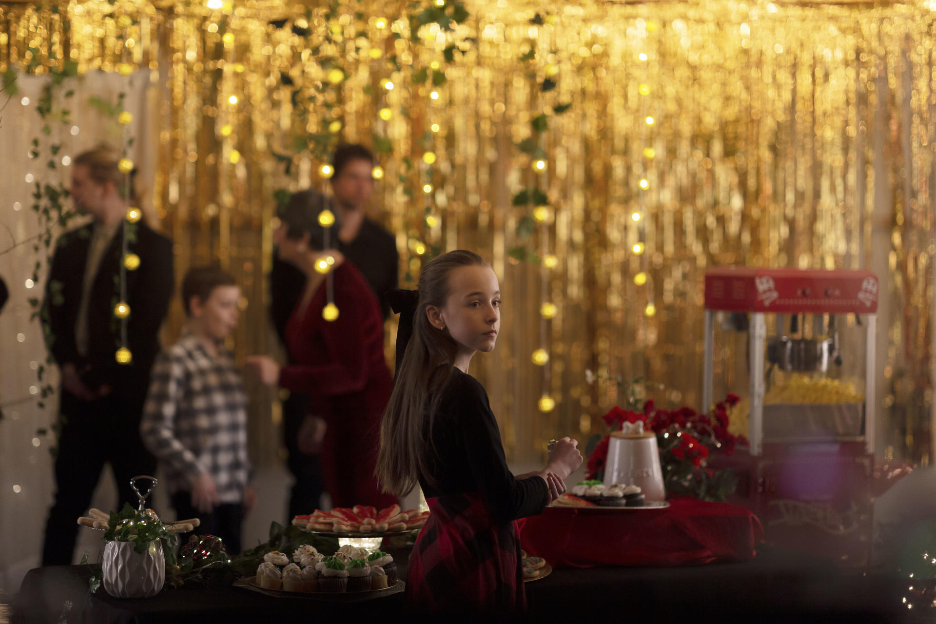 Ein Mädchen in einem karierten Kleid blickt auf einer Party zurück zum Betrachter. Goldene Luftschlangen und eine Popcornmaschine sind im Hintergrund.