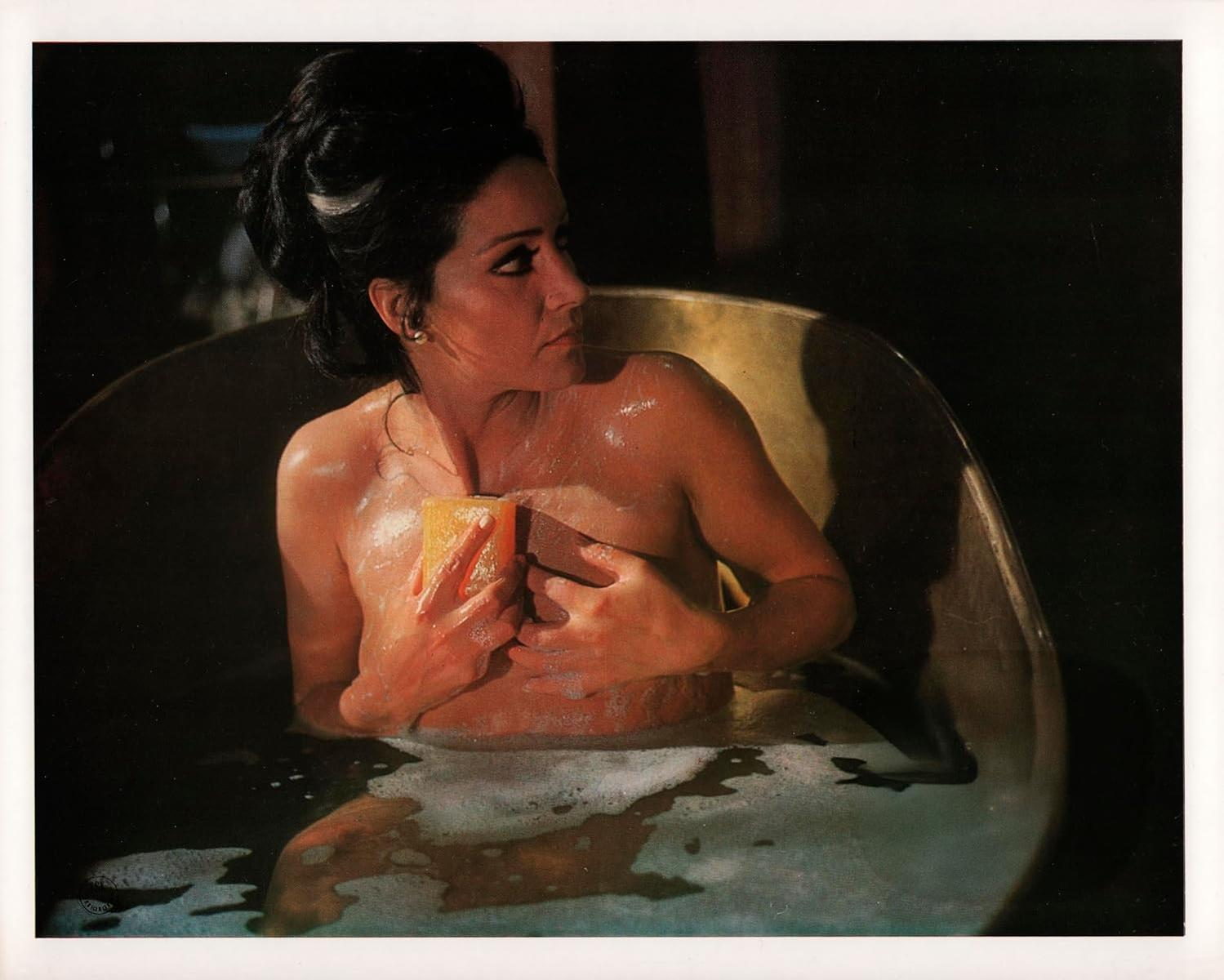 Eine Frau in einer Badewanne hält einen gelben Schwamm und bedeckt ihre Brust. Die Szene ist schwach beleuchtet, mit Blasen und einem Vintage-Feeling.