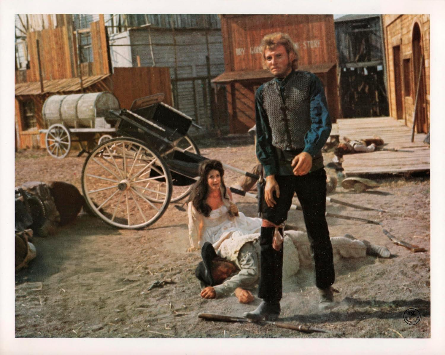 Eine Western-Filmszene. Ein Mann mit Weste steht über einem Mann am Boden. Eine Frau sitzt in der Nähe. Wagen und Gebäude sind im Hintergrund.