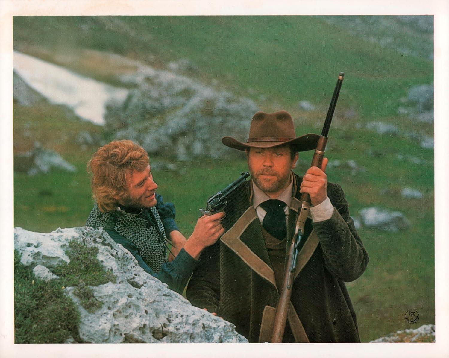 Zwei Männer in Bergkulisse, einer mit Gewehr und Cowboyhut, der andere mit einer Waffe.