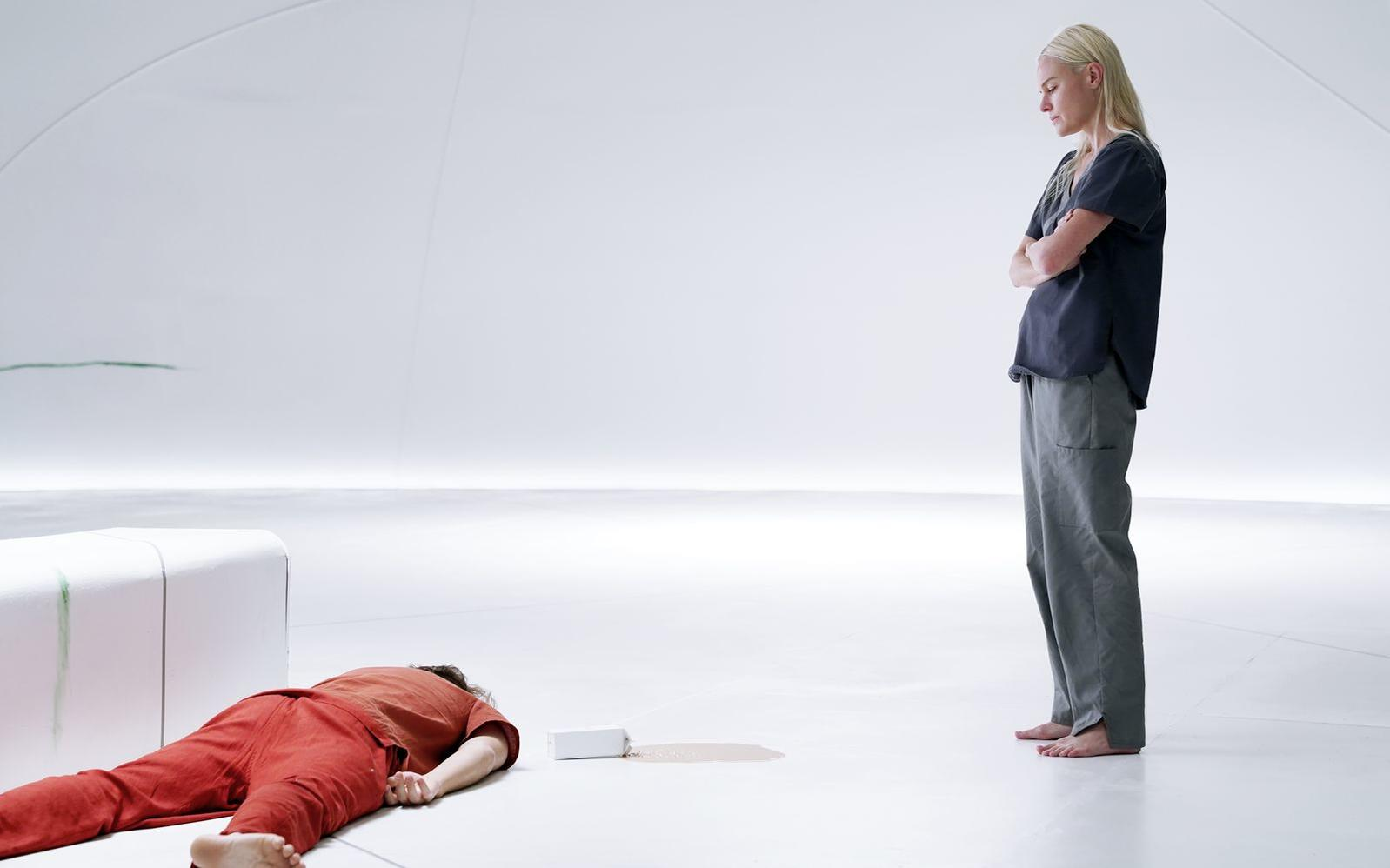 Eine Frau steht und blickt auf eine Person, die auf dem Boden liegt. Die Person trägt eine rote Hose und ein braunes Hemd. Daneben befindet sich eine verschüttete Flüssigkeit.