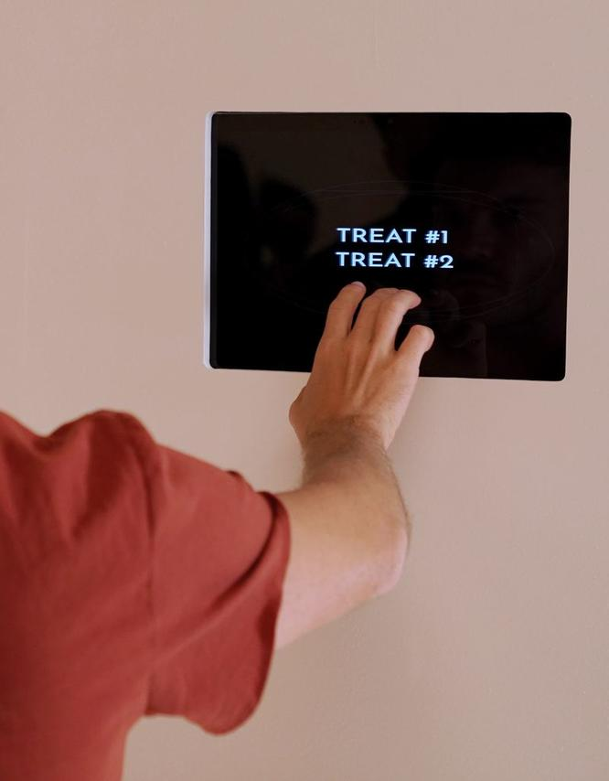 Eine Hand berührt einen schwarzen Bildschirm mit "TREAT #1" und "TREAT #2". Die Person trägt ein rotes Hemd.