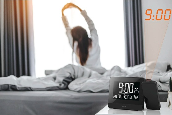 Osoba rozciąga się w łóżku. Zegar cyfrowy pokazuje 9:00. Czas jest wyświetlany na ścianie. Scena to sypialnia.
