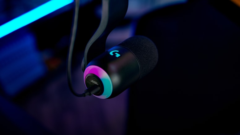 Egy fekete mikrofon világító Logitech 'G' logóval. A talpán színes gyűrű van. A háttér sötét.