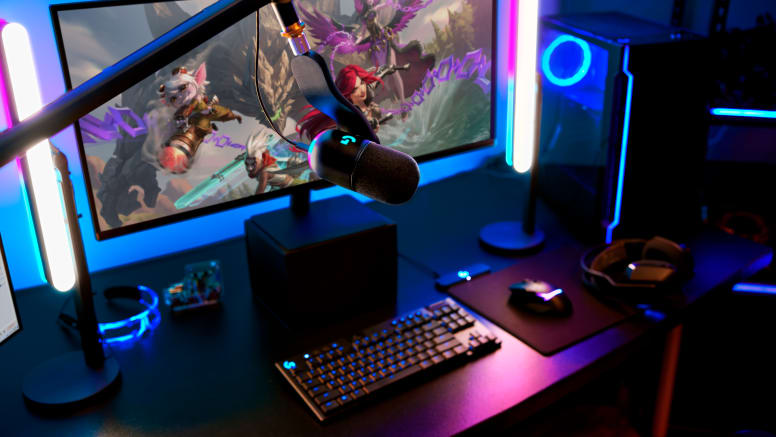 Játékbeállítás monitorral, ami játékot mutat, mikrofon, billentyűzet, egér, számítógép és LED-lámpák. Sötét szoba kék és rózsaszín világítással.