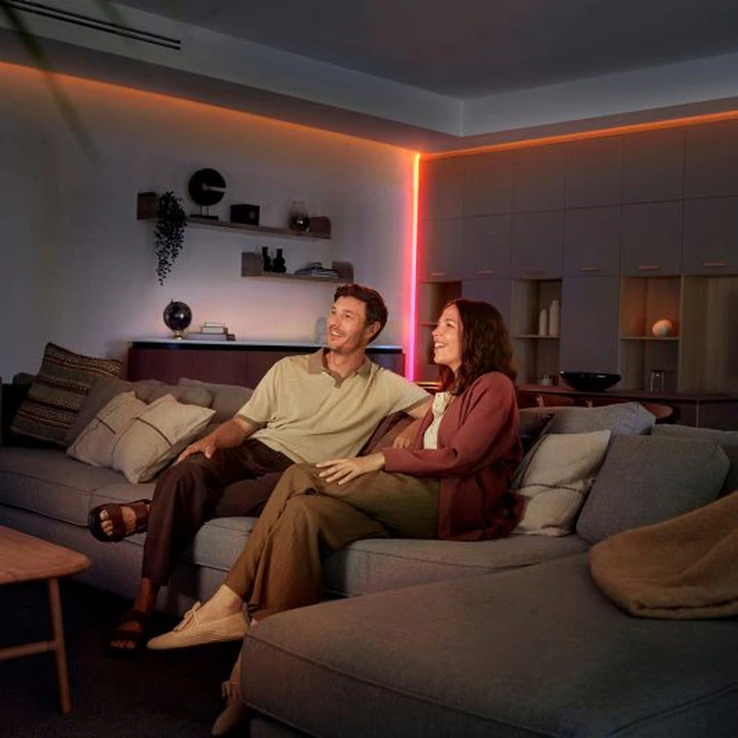 A couple sits on a gray sofa, smiling. A warm orange light is on the ceiling. A wooden table is in front of them.