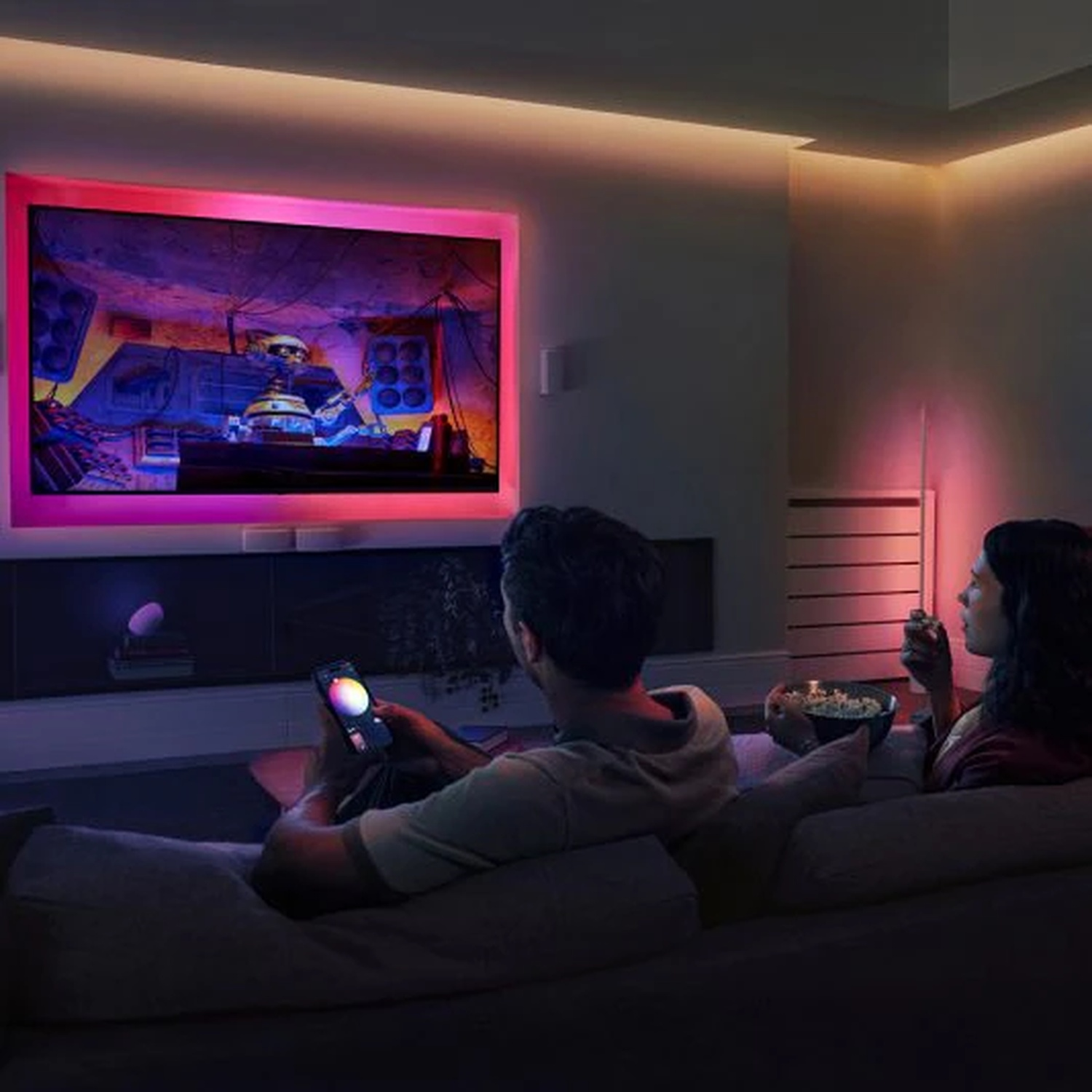 Couple on a couch watching TV with colorful lights. Man holds a phone, woman eats popcorn. TV has bright pink frame.