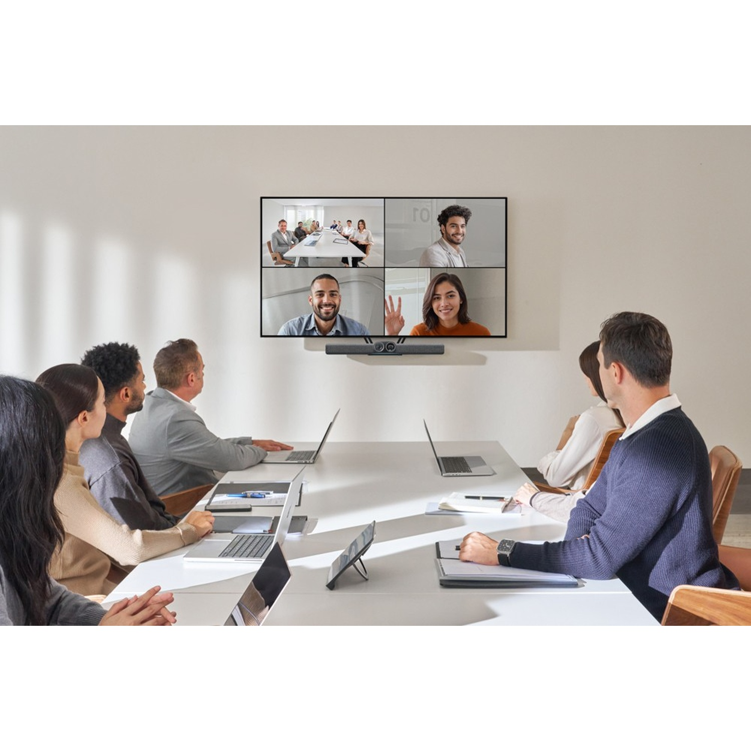 Un gruppo di persone in una riunione, alcune su laptop. Uno schermo mostra una videoconferenza con quattro persone.