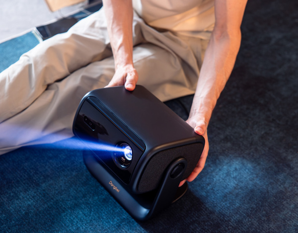 Osoba trzyma czarny projektor, który emituje niebieską wiązkę. Osoba siedzi na ciemnoniebieskim dywanie, widoczne nogi.