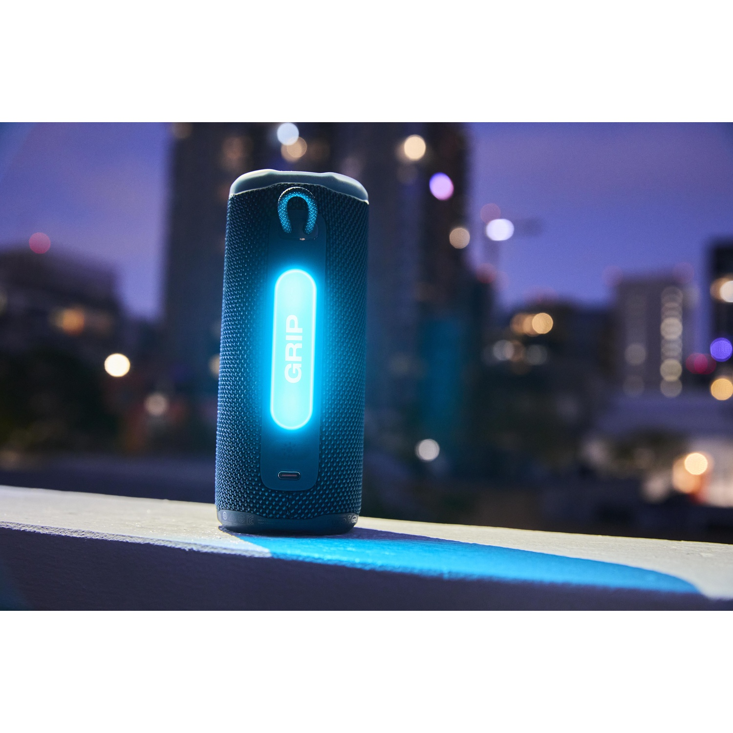 Un altoparlante 'GRIP' nero con luce blu, posizionato su un davanzale. Edifici della città sullo sfondo con luci sfocate, al crepuscolo.