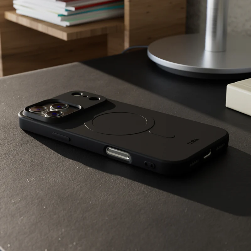 Ein schwarzes iPhone liegt auf einem Schreibtisch. Eine Handyhülle mit kreisförmigem MagSafe-Design ist sichtbar. Im Hintergrund befinden sich eine Lampe und ein Regal.