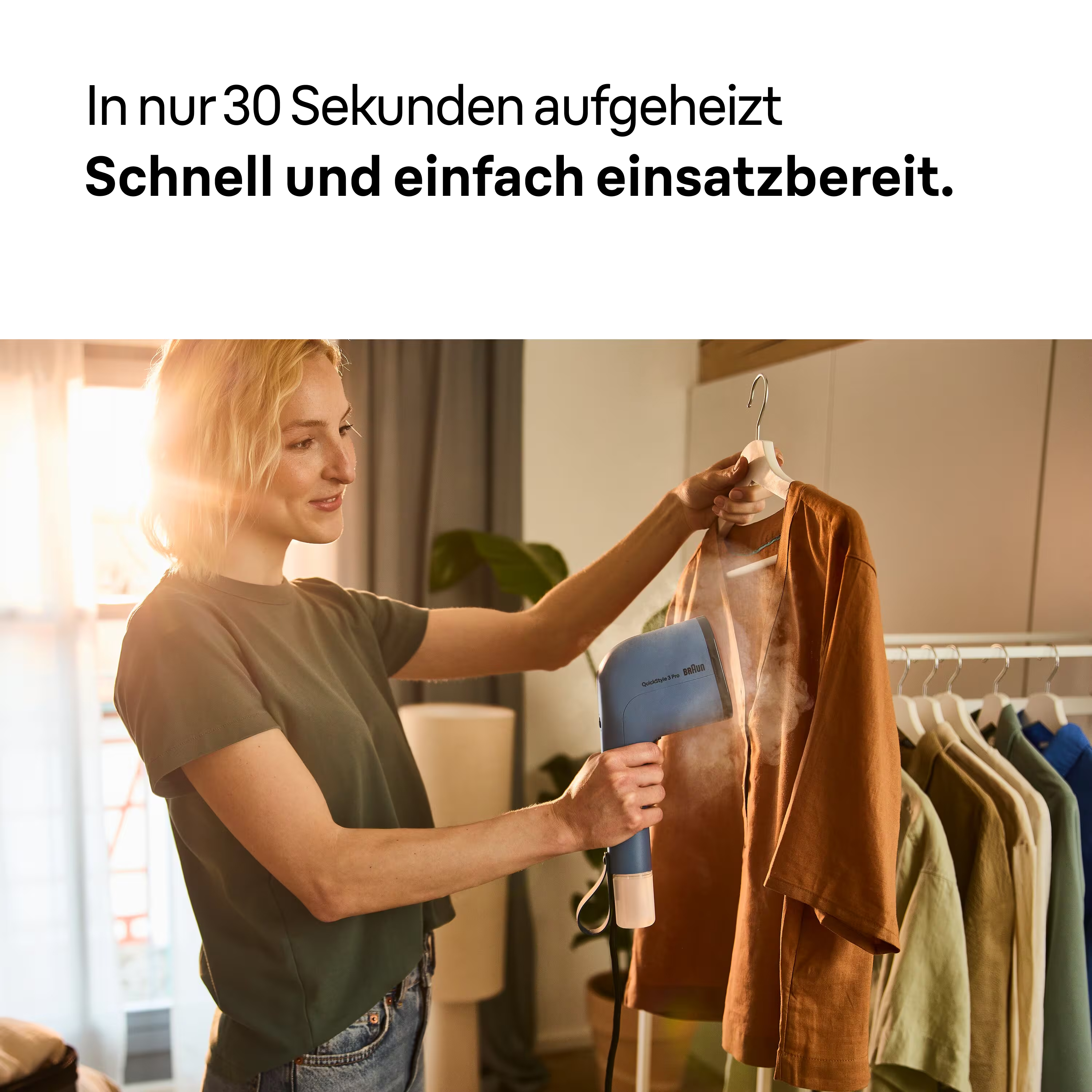 Eine Frau bügelt ein braunes Hemd mit einem blauen Steamer. Kleidung hängt im Hintergrund auf einem Gestell.