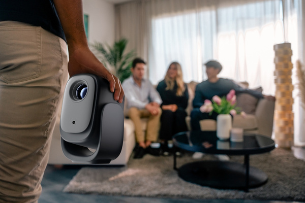 Osoba trzyma czarno-szary projektor. Trzy osoby siedzą na kanapie w tle. Stolik kawowy jest na pierwszym planie.