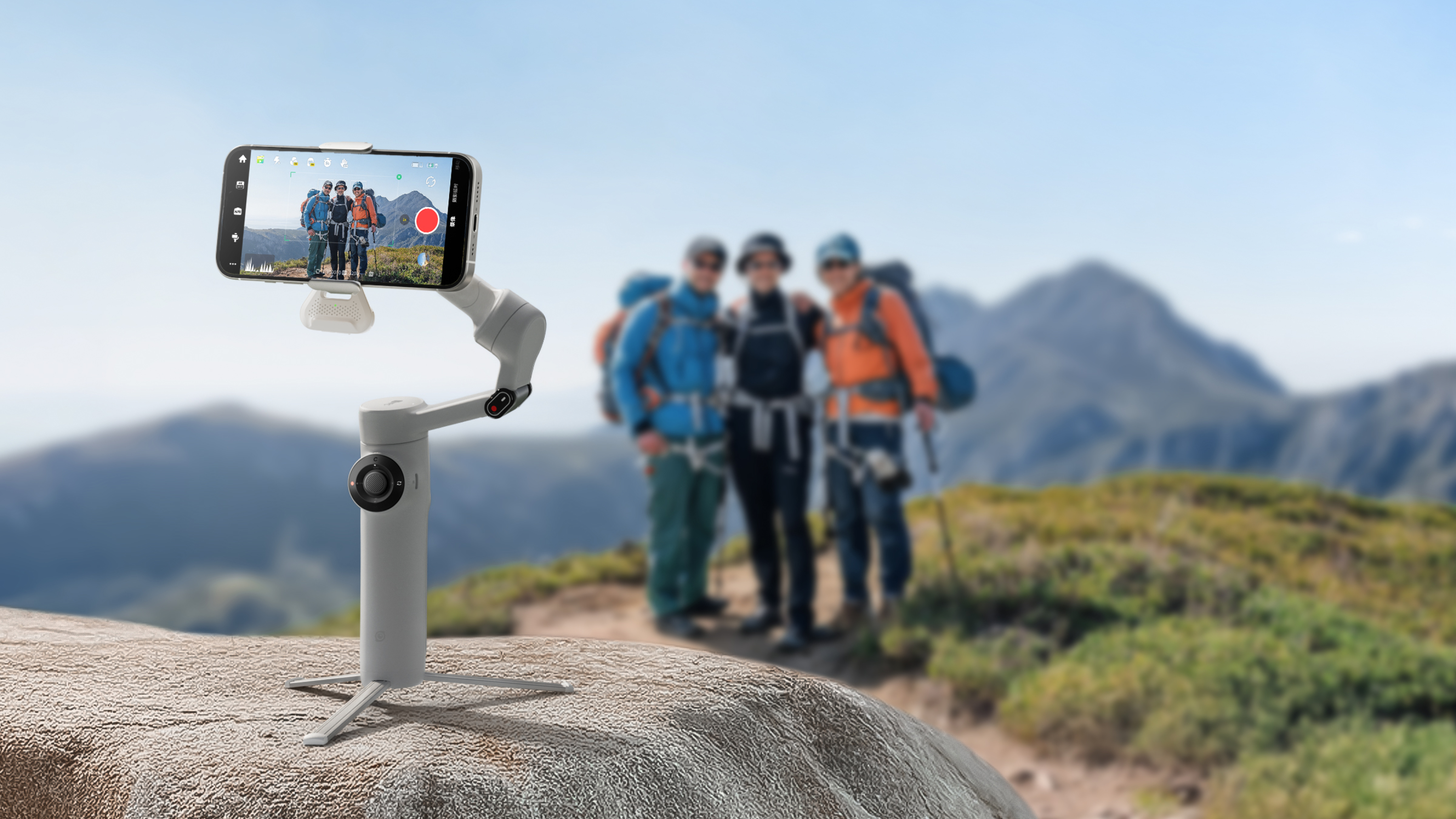 Ein Telefon auf einem Gimbal nimmt Wanderer in den Bergen auf. Das Telefon zeigt drei Wanderer. Der Hintergrund hat Berge und blauen Himmel.