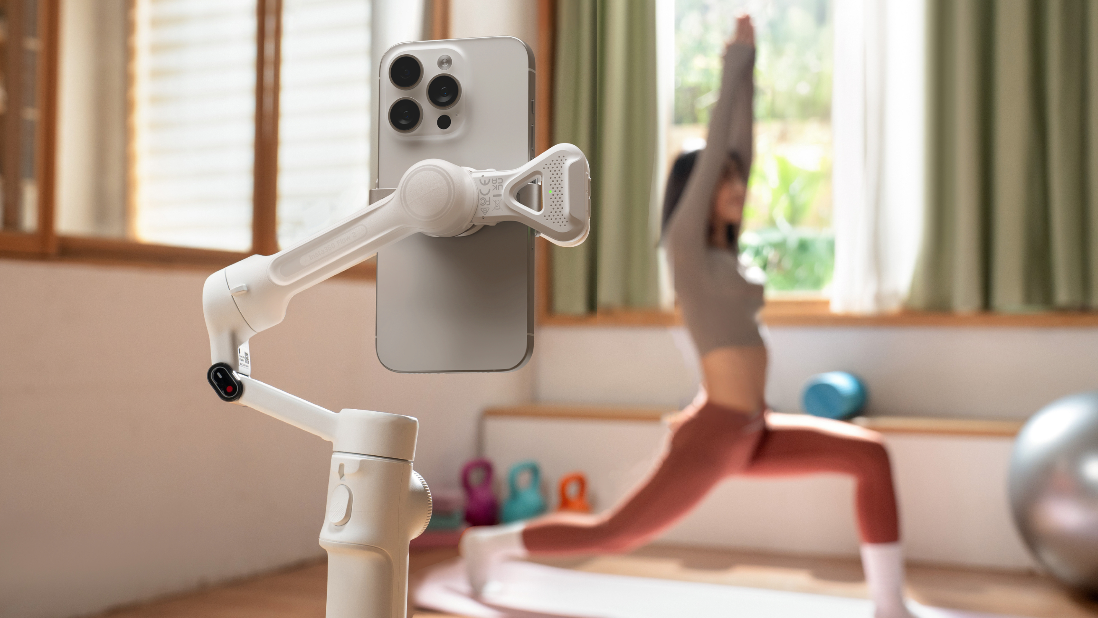 Ein Telefon auf einem weißen Gimbal, das eine Person beim Yoga filmt. Der Hintergrund umfasst Fenster und grüne Vorhänge.