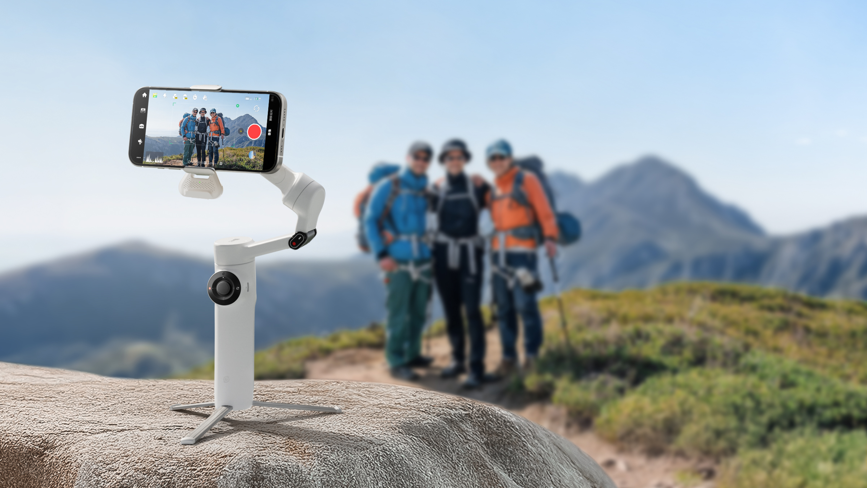 Ein Telefon auf einem Gimbal zeichnet Wanderer in den Bergen auf. Das Gimbal steht auf einem Felsen, der Hintergrund zeigt Berge und Himmel.