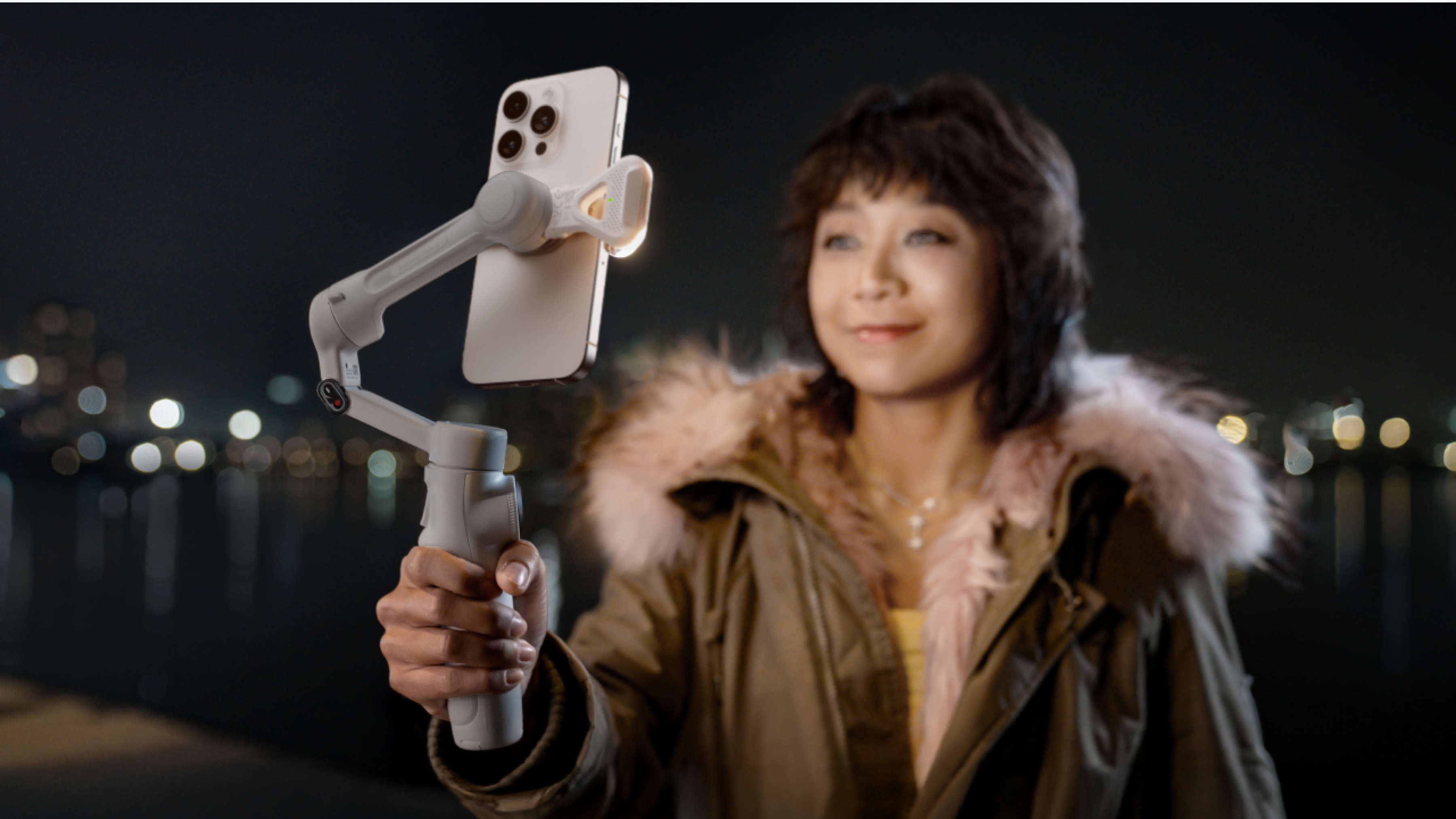 Eine Frau hält nachts ein Telefon auf einem Gimbal. Sie trägt einen Mantel mit Pelzkragen, der Hintergrund hat Bokeh-Lichter.