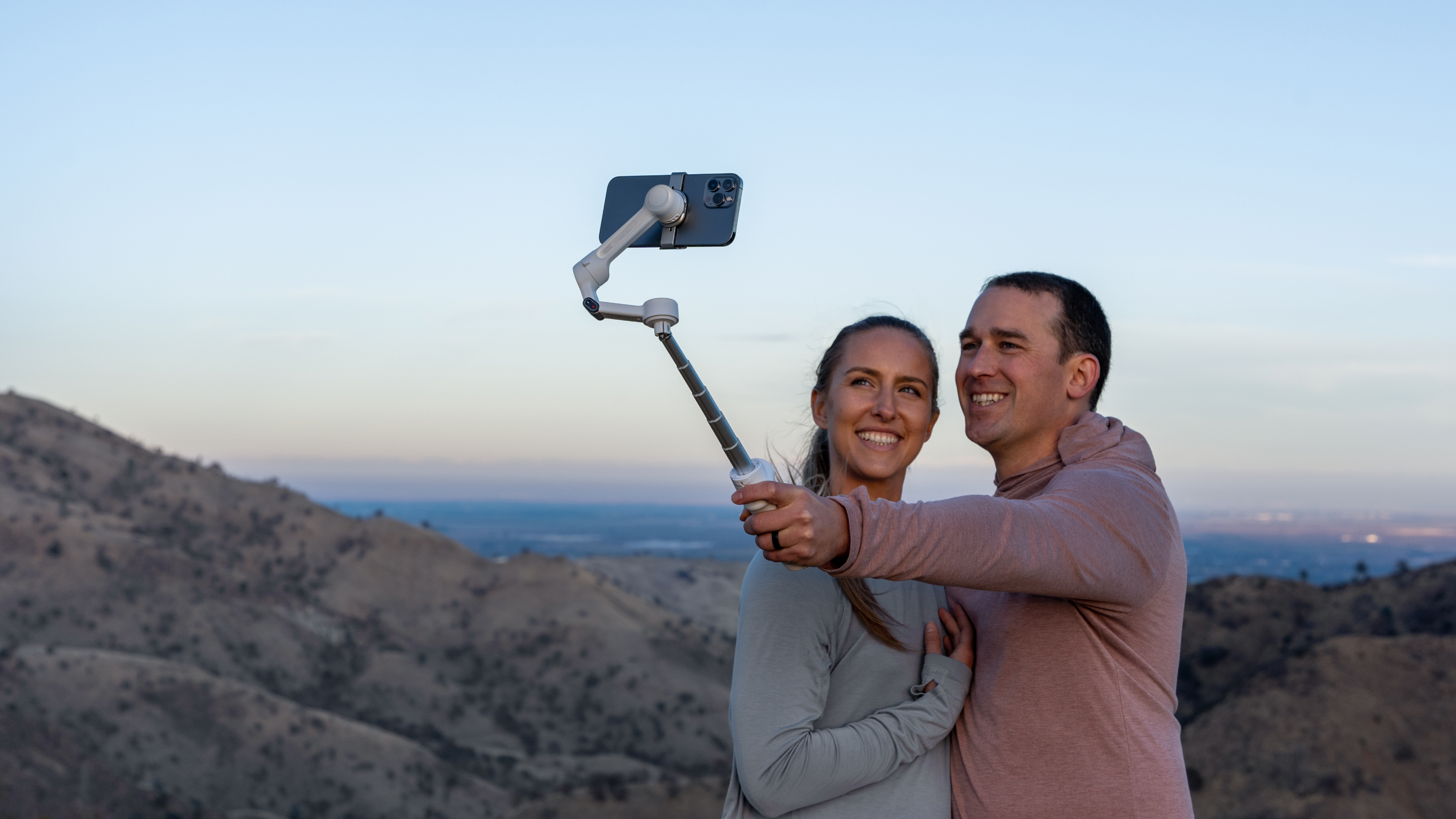 Ein Paar lächelt, während es mit einem Handy auf einem Selfie-Stick Selfies macht, mit Bergen im Hintergrund.