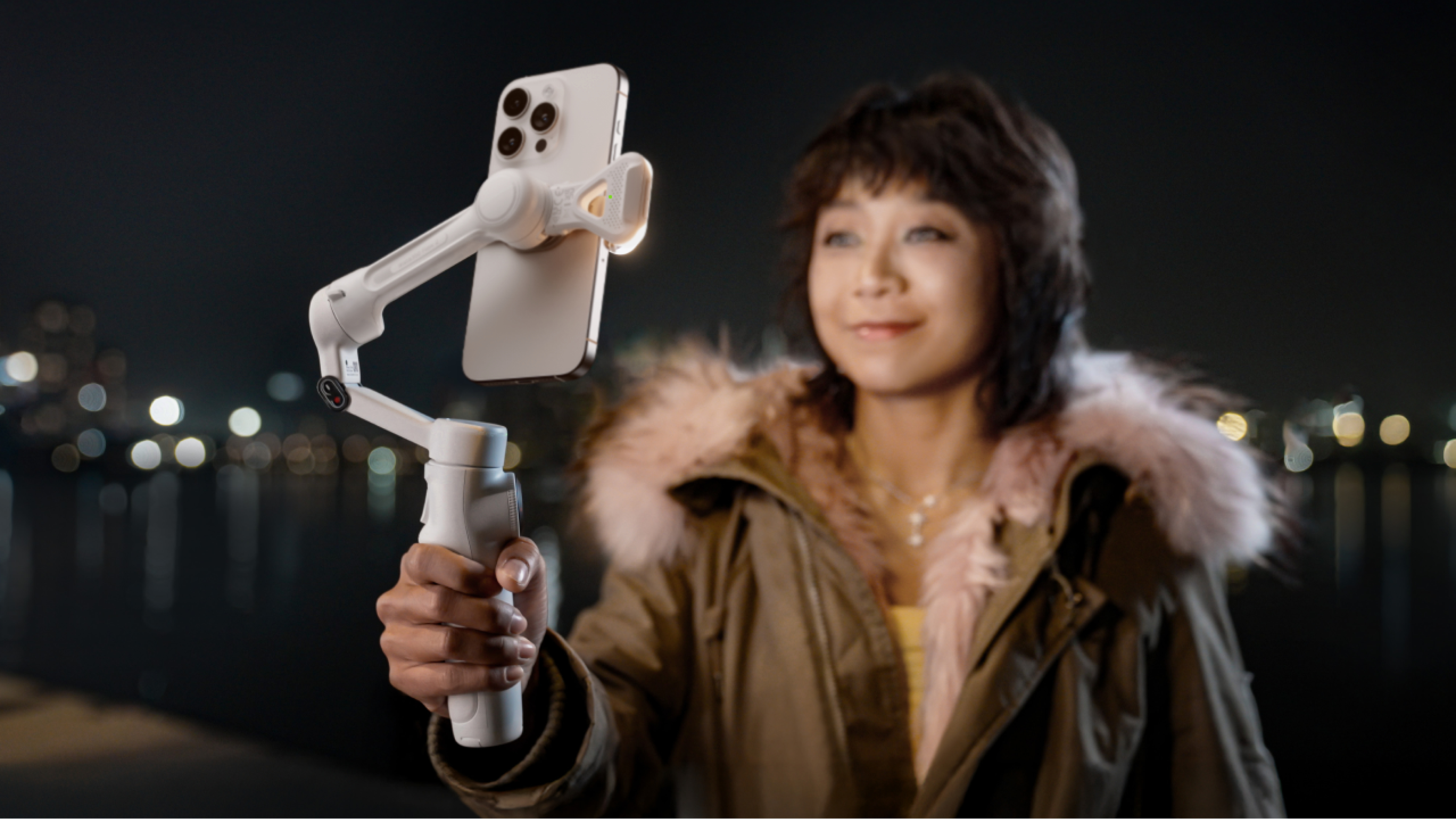 Eine Frau hält ein Telefon auf einem Gimbal. Sie lächelt leicht. Der Hintergrund hat Lichter und ein Gewässer.