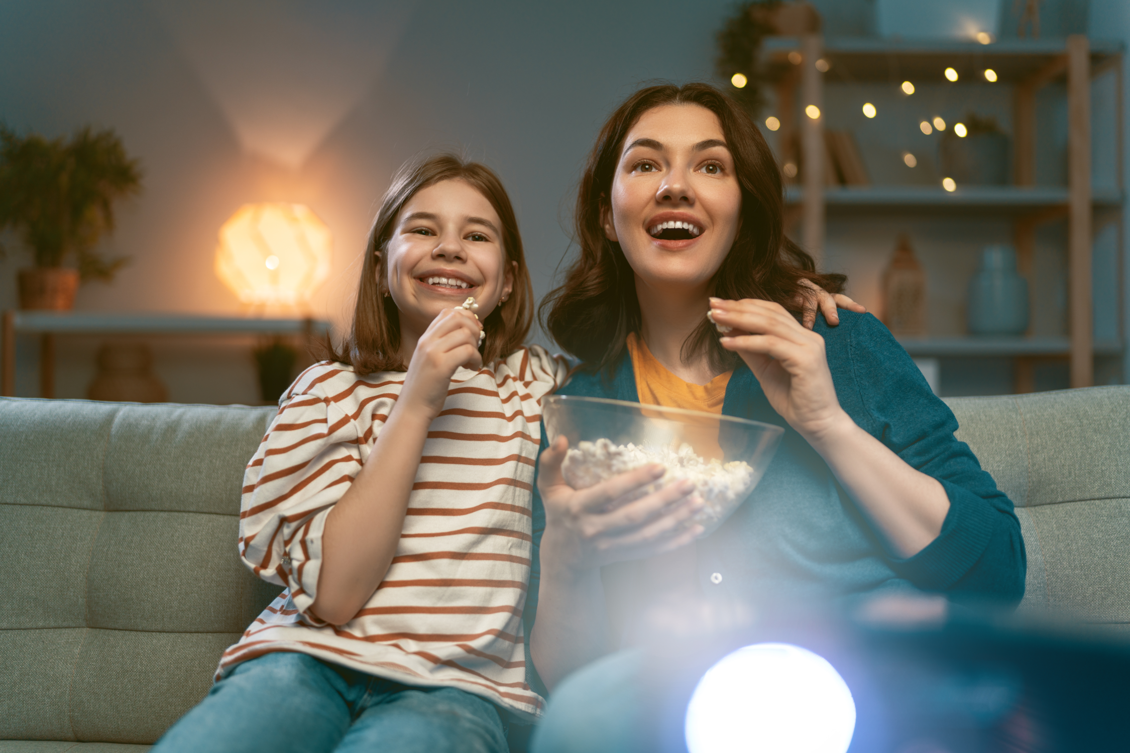 Une femme et une fille sont assises sur un canapé, mangeant du pop-corn. Elles regardent en haut et sourient. Un projecteur brille au premier plan.