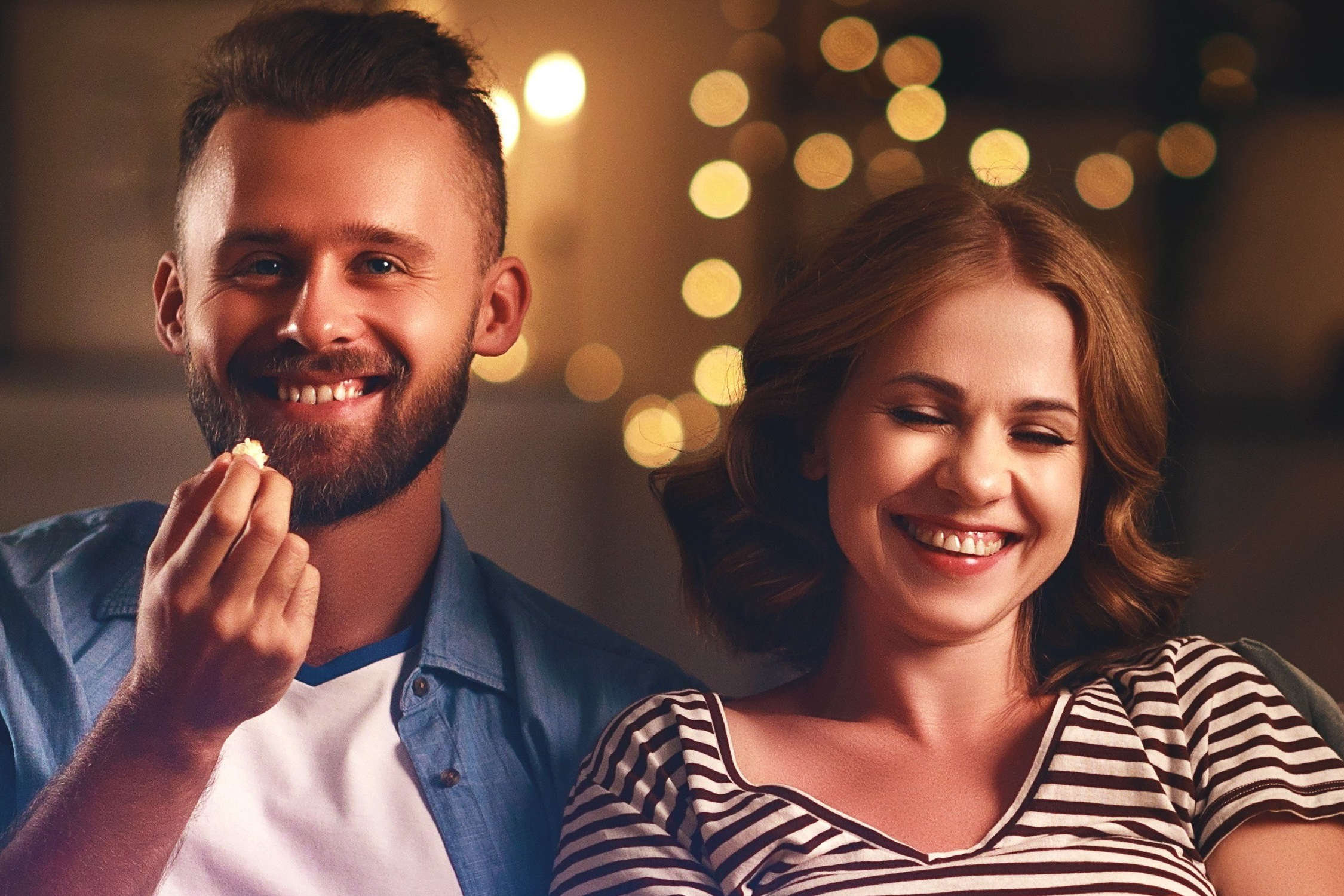 Un couple souriant, l'homme tient du pop-corn. Les yeux de la femme fermés, souriante. Fond: lumières floues.