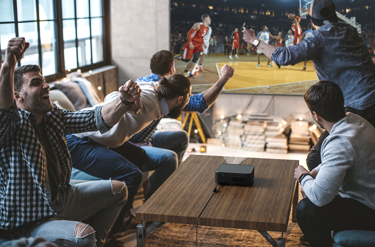 Männer schauen Basketball auf einem Bildschirm. Sie jubeln mit erhobenen Armen. Ein Projektor steht auf einem Tisch. Im Hintergrund sind Fenster und Stadion zu sehen.