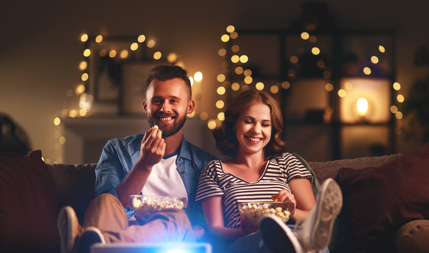 Ein Paar sitzt auf einem Sofa, isst Popcorn und lächelt. Warme Beleuchtung und eine Projektionsfläche sind sichtbar.
