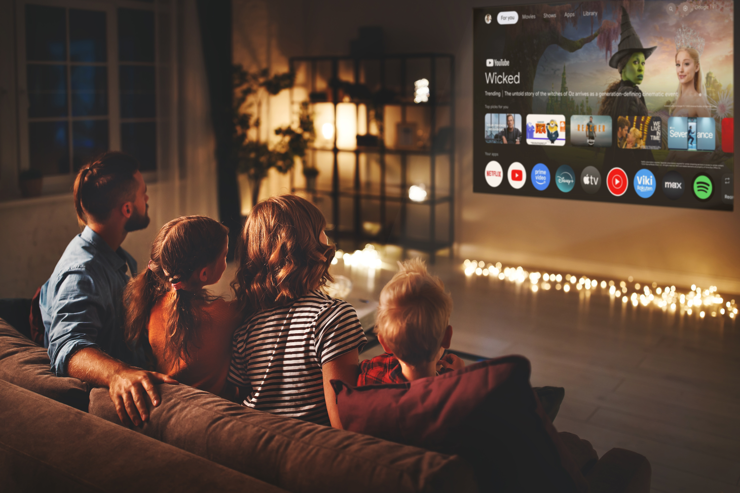 Eine Familie sieht fern. Auf dem Fernseher läuft 'Wicked' auf YouTube. Sie sitzen auf einem Sofa in einem warm beleuchteten Wohnzimmer.