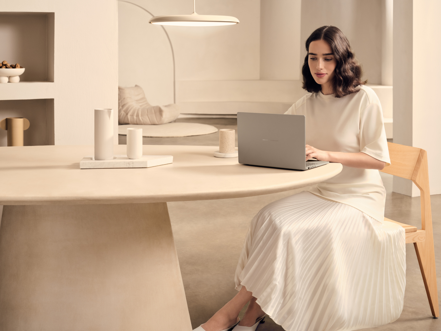 Mujer sentada en una mesa usando una laptop. Lleva un vestido blanco. La habitación es moderna, con una mesa redonda de color claro.